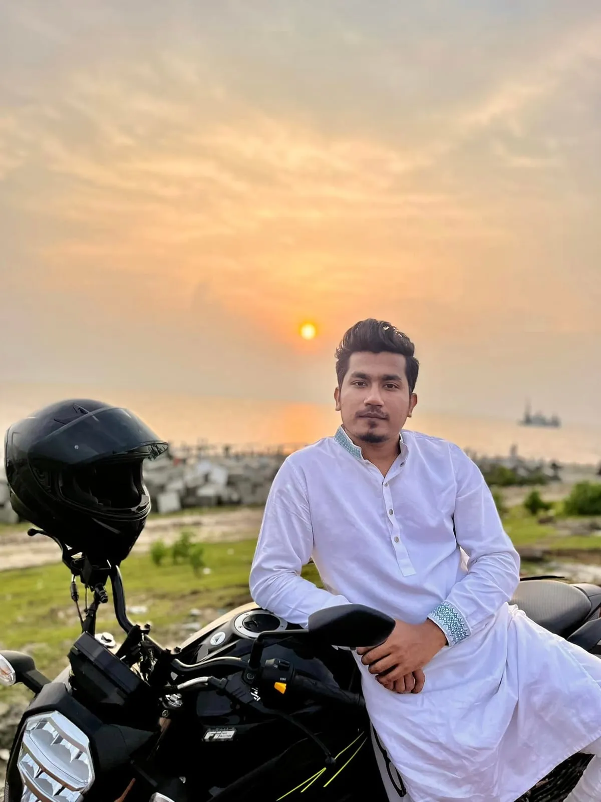 Shakawat Hridoy in a white panjabi sitting on a motorcycle at sunset by the water