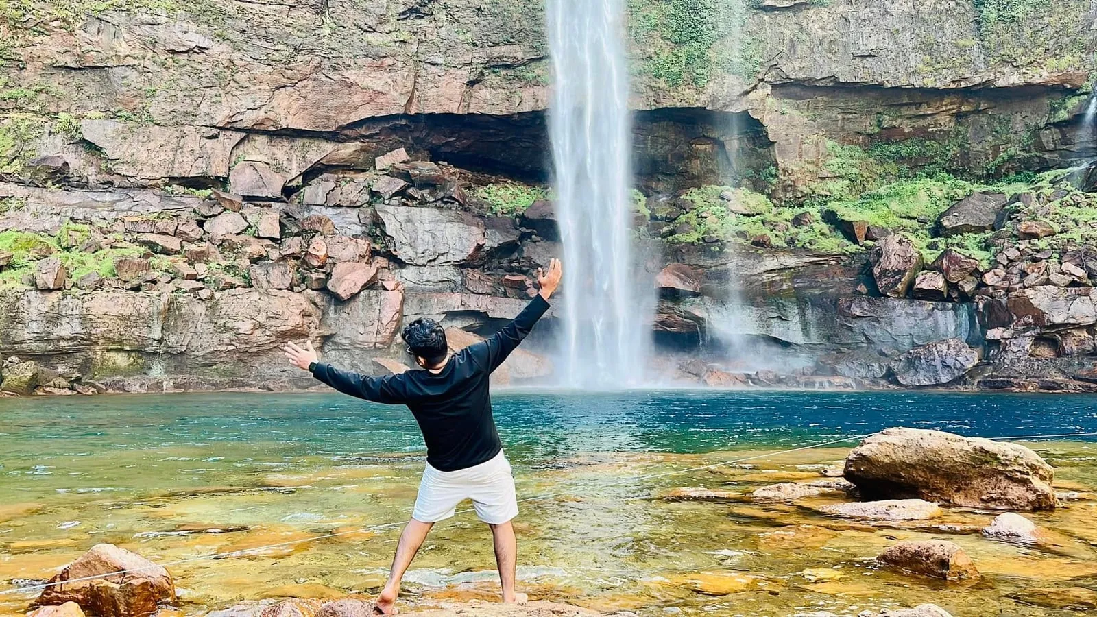 Shakawat Hridoy standing with his arms wide in the pool below a tall waterfall in Meghalaya, India