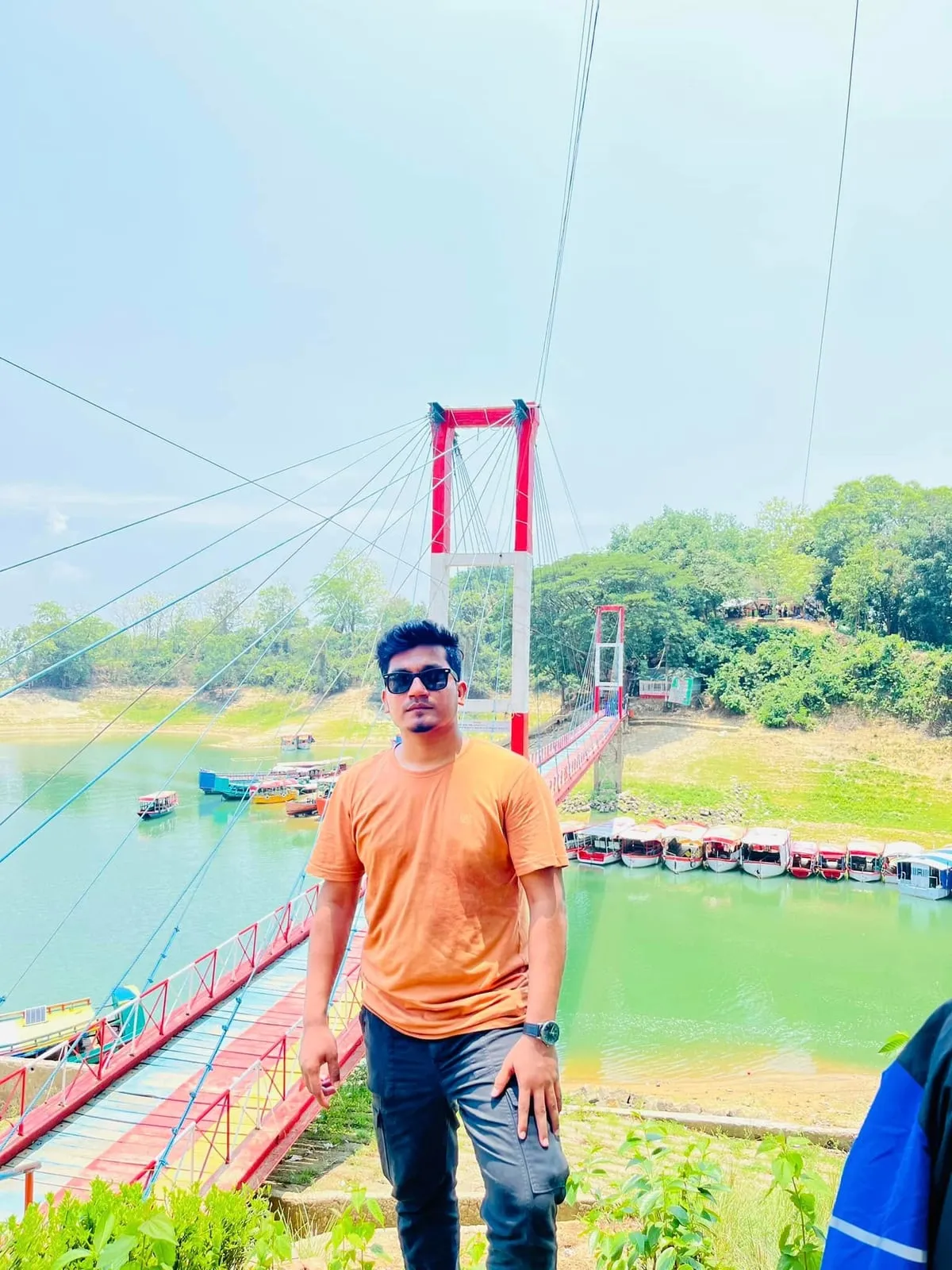 Shakawat Hridoy in an orange t-shirt in front of the red hanging bridge over the lake in Rangamati