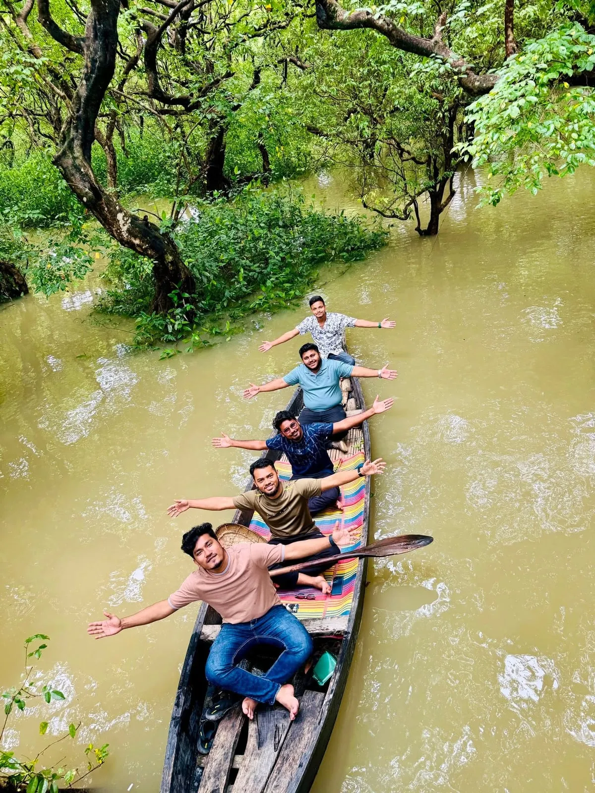 Shakawat Hridoy and friends with their arms out in a long wooden boat in the flooded Ratargul swamp forest, Sylhet